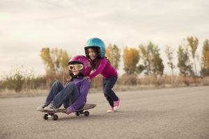 Two young girls are racing on a skateboard. One girl is pushing the other and they are going really fast on a rural road. They are young adventurers wearing helmets and flight goggles and are ready to beat all the boys in the race. Image taken in Utah, USA.